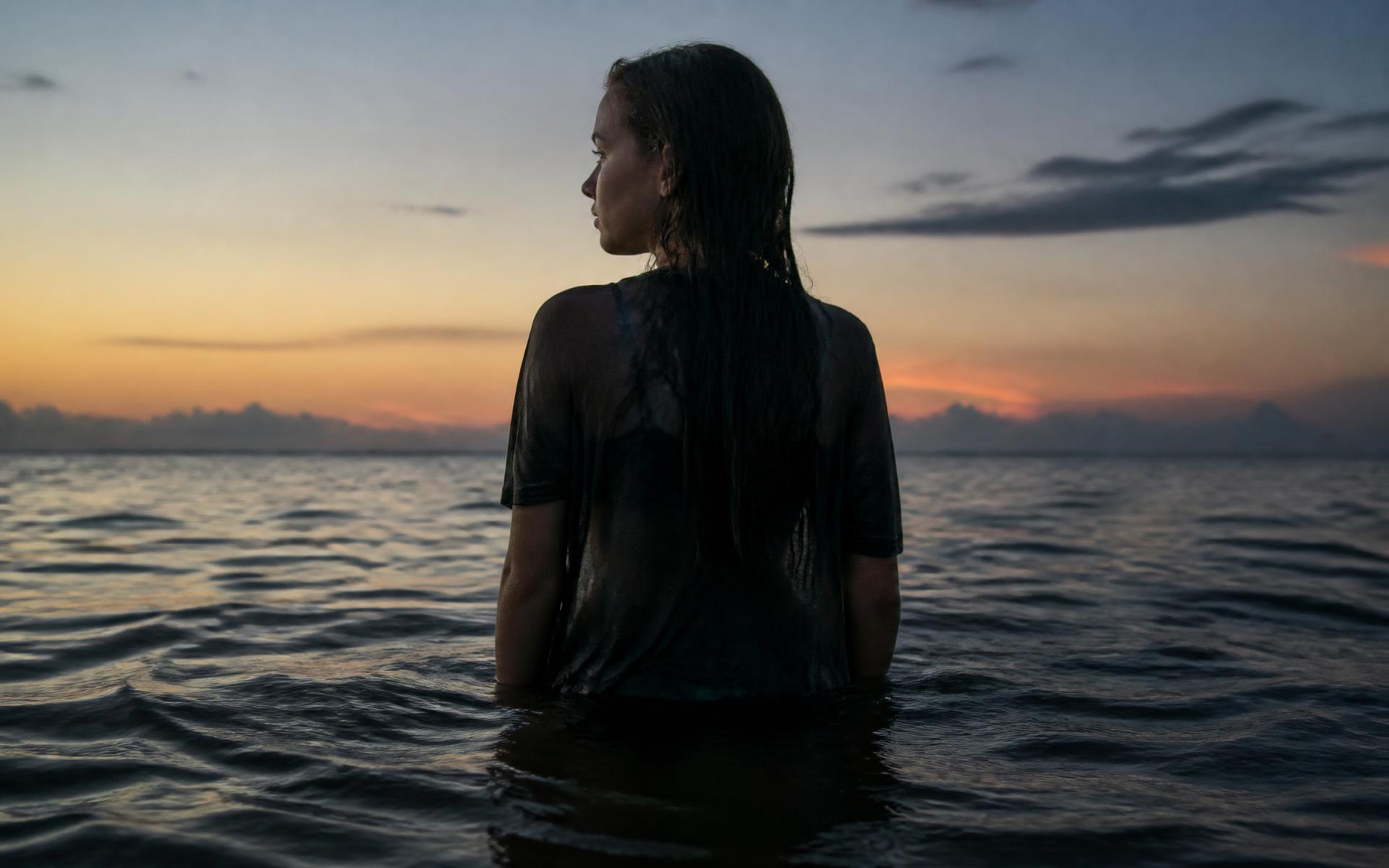 A young woman standing waist deep in the sea, looking towards the horizon at dusk